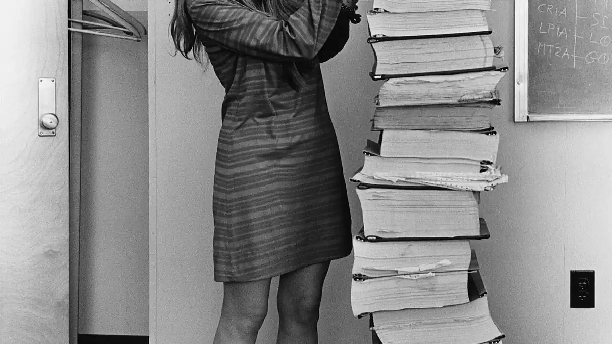 Margaret Hamilton standing in 1969 beside towering vertical stacks of Apollo 11 guidance software listings.