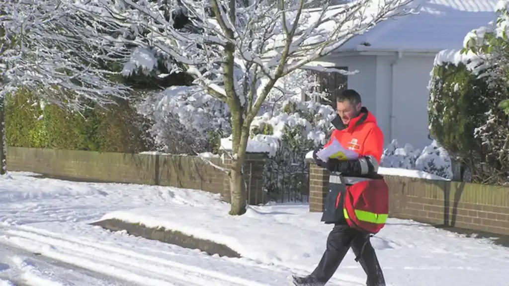 A uniformed Royal Mail postman carrying a red mail delivery bag along a residential street.