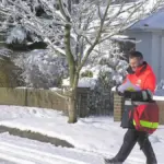 A uniformed Royal Mail postman carrying a red mail delivery bag along a residential street.