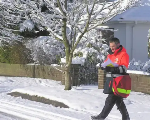 A uniformed Royal Mail postman carrying a red mail delivery bag along a residential street.