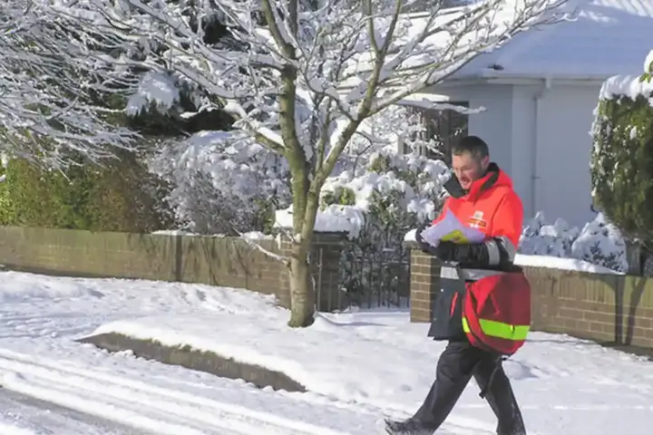 A uniformed Royal Mail postman carrying a red mail delivery bag along a residential street.