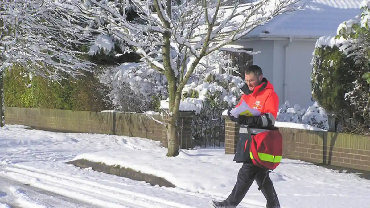 A uniformed Royal Mail postman carrying a red mail delivery bag along a residential street.