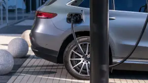 Rear quarter of a silver electric car connected to a public charging station beside modern urban paving.