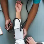 Blood pressure monitoring gauge and inflatable arm cuff resting on a wooden examination table in a clinical setting.