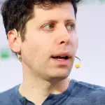 OpenAI CEO Sam Altman speaking on stage in a dark shirt against a blue event backdrop.
