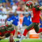 American football player attempts a field goal during a game, with teammates and sideline personnel visible behind him.