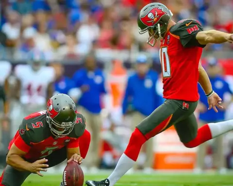 American football player attempts a field goal during a game, with teammates and sideline personnel visible behind him.