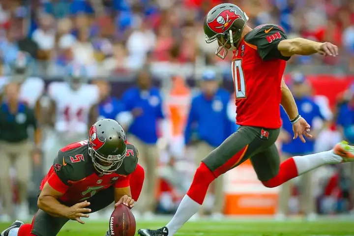 American football player attempts a field goal during a game, with teammates and sideline personnel visible behind him.