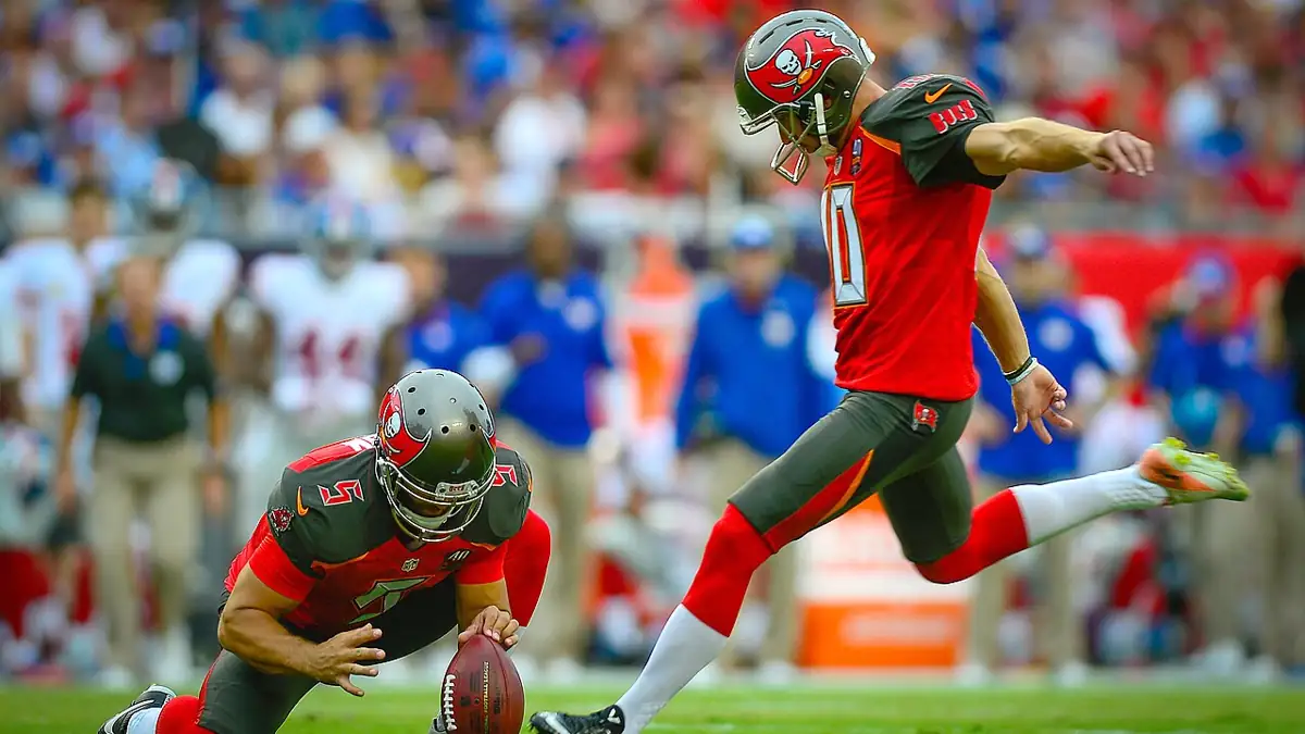 American football player attempts a field goal during a game, with teammates and sideline personnel visible behind him.