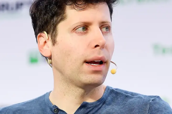 OpenAI CEO Sam Altman speaking on stage in a dark shirt against a blue event backdrop.