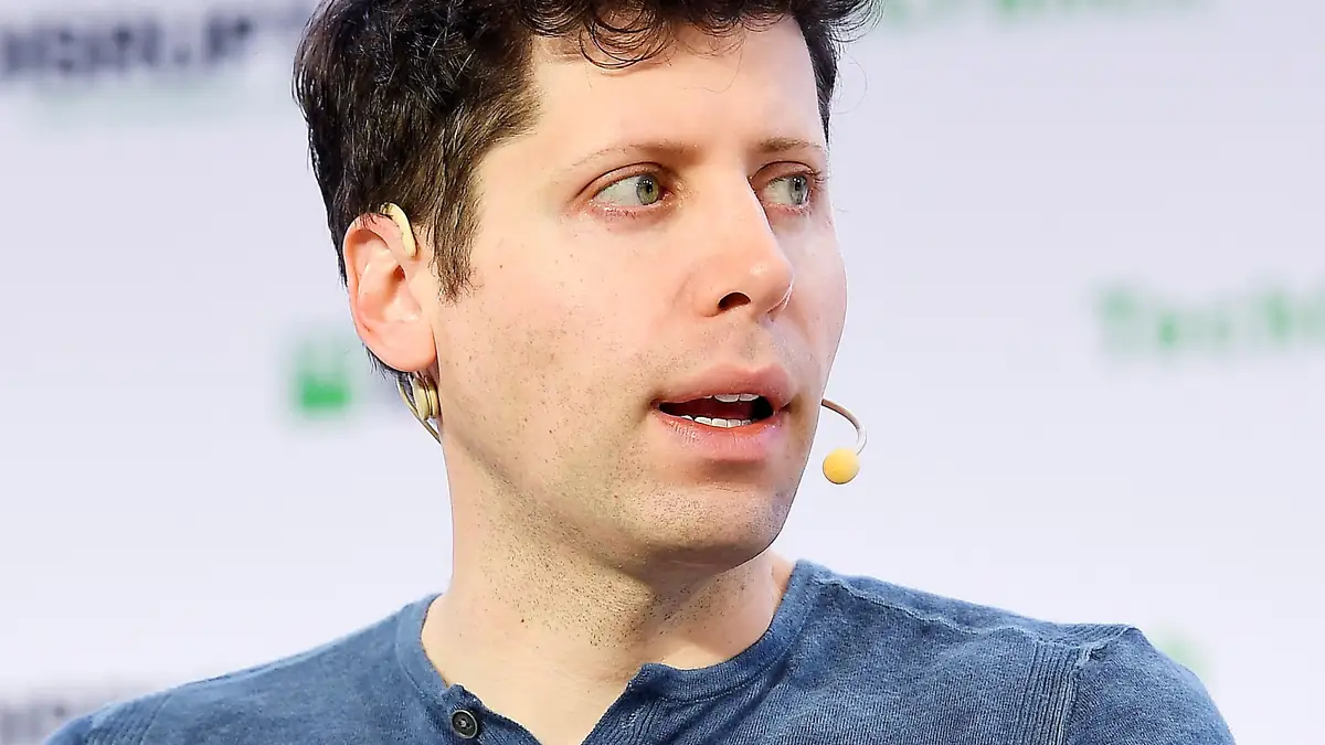 OpenAI CEO Sam Altman speaking on stage in a dark shirt against a blue event backdrop.