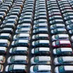 Rows of new cars parked in a large outdoor inventory lot viewed from above.