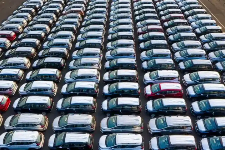 Rows of new cars parked in a large outdoor inventory lot viewed from above.