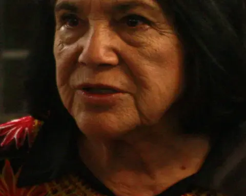 Close portrait of labor organizer Dolores Huerta speaking at a public event with a microphone and audience behind her.