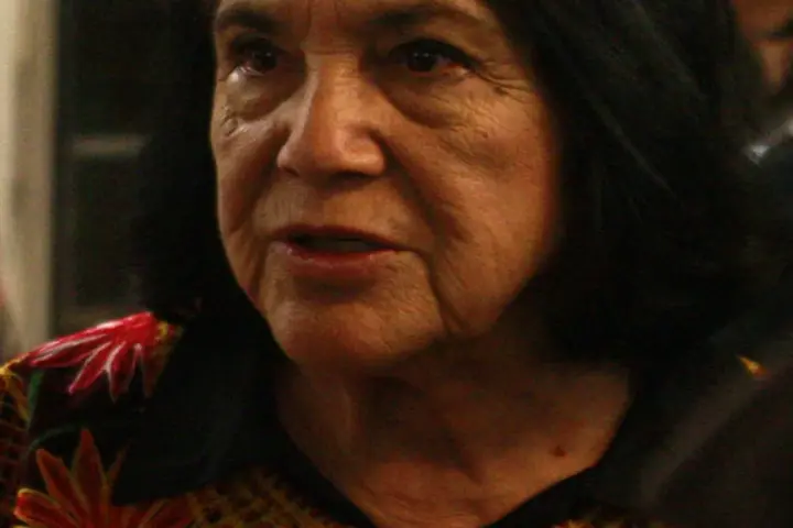 Close portrait of labor organizer Dolores Huerta speaking at a public event with a microphone and audience behind her.