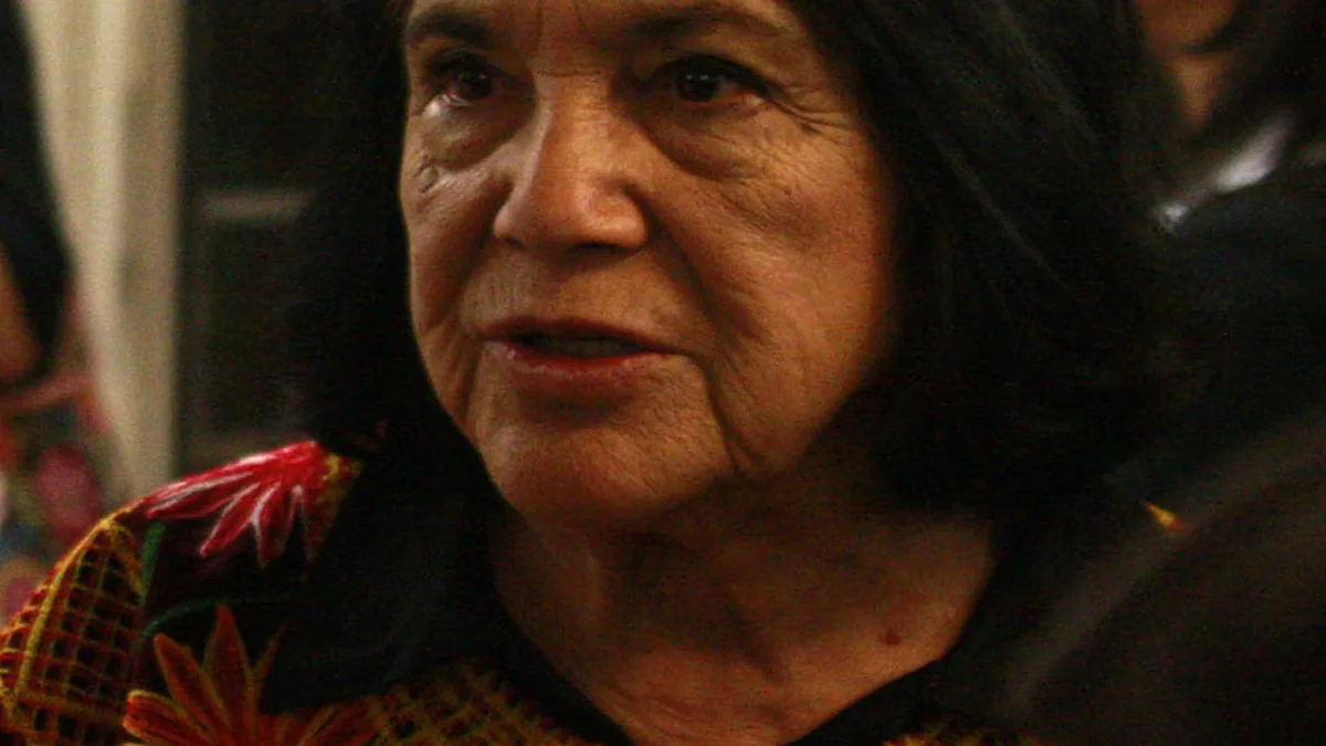 Close portrait of labor organizer Dolores Huerta speaking at a public event with a microphone and audience behind her.