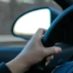 Close-up of a driver's hand gripping a steering wheel while driving, with blurred dashboard controls visible