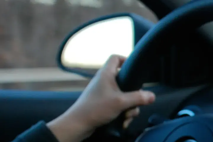 Close-up of a driver's hand gripping a steering wheel while driving, with blurred dashboard controls visible