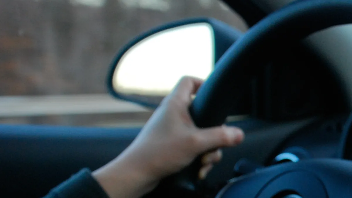 Close-up of a driver's hand gripping a steering wheel while driving, with blurred dashboard controls visible